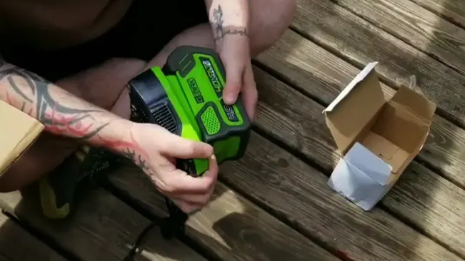person with tattoos on their arms, using a green and black power tool near an open cardboard box on a wooden surface, likely outdoors.