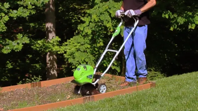 Individual operating a green tiller in a raised garden bed on a sunny day