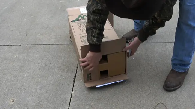 Person in camouflage jacket and jeans picking up a cardboard box outdoors