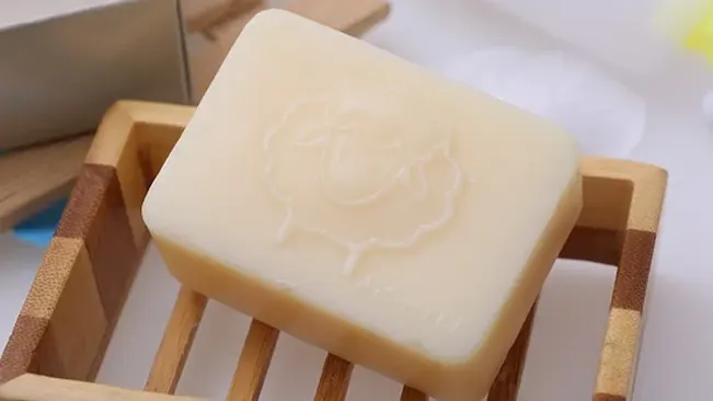 white bar of soap with a rose imprint, resting on a square-shaped wooden soap dish