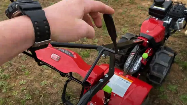 shows a close-up of a person’s hand adjusting the handle of a red garden tiller on a grassy ground