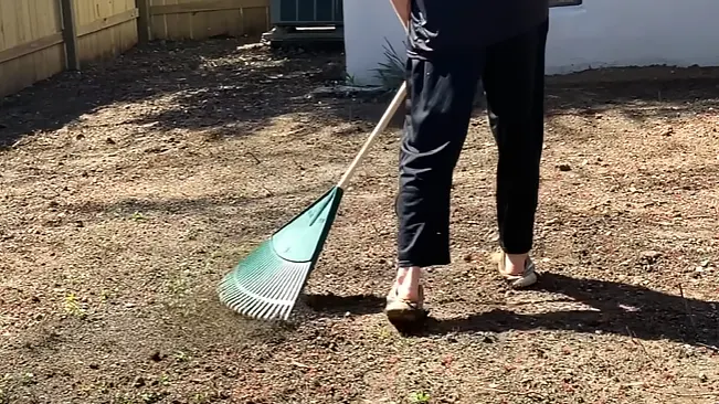 A person in black pants and tan shoes raking over a patch of bare earth in a sunny yard.