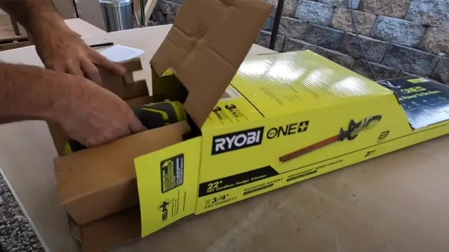 Person unboxing a new RYOBI ONE+ hedge trimmer on a flat surface with a stone wall in the background