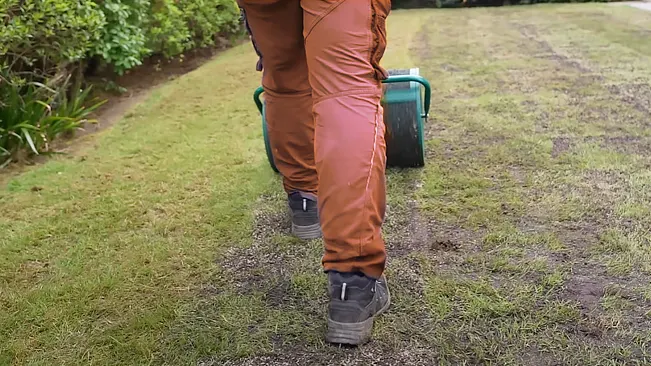 A person in brown trousers pushing a green wheelbarrow across a grassy lawn.