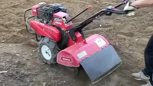 A red Honda tiller being operated on bare soil by a person partially visible on the right side of the frame.