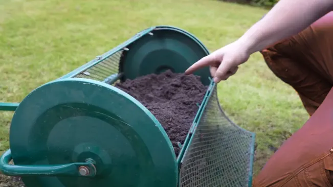A person's hand pointing to a wheelbarrow filled with dark, rich soil.