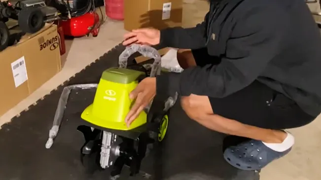 a person, visible from the waist down, assembling a small green machine with black wheels and metallic components on the floor of a garage