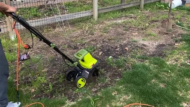 person operating a small, green and black tiller on a garden bed surrounded by a wooden fence