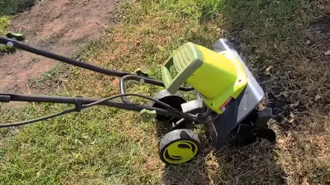a green and black lawn mower on a patch of grass and soil, casting a shadow under sunlight