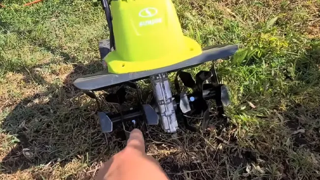 person’s finger pointing at a bright green garden tiller with the brand logo “SUN JOE” on a grassy surface