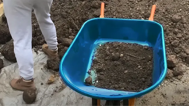 A blue wheelbarrow filled with soil and rocks, with a person in white pants and brown boots standing beside it on uneven ground.