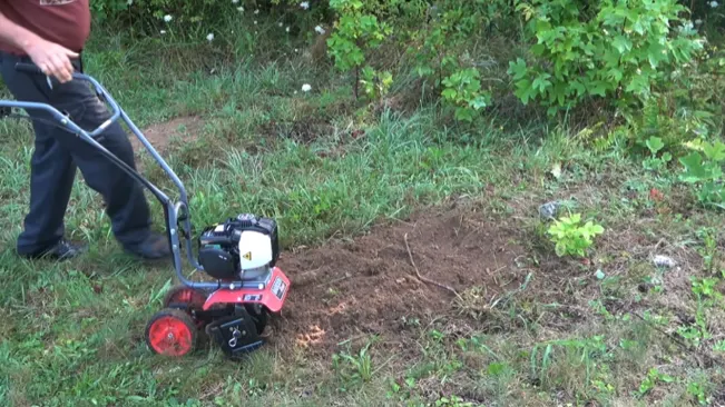 person using a red and black rototiller in a garden, preparing the soil for planting. The background features greenery including grass and bushes.