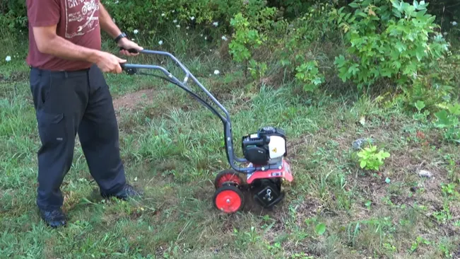 a person operating a small, red and black rototiller on uneven grassy ground in a garden.