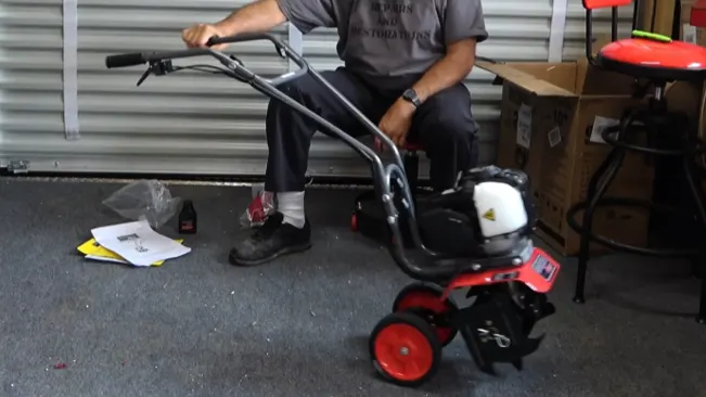 a person seated in a garage, assembling a small red and black tiller with three wheels.