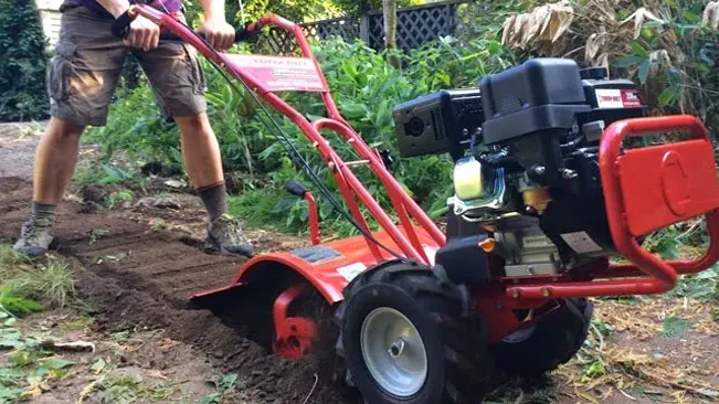 Person operating a red garden tiller on soil amidst greenery