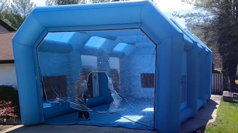 A person stands inside a large blue inflatable paint booth with transparent sections and an open doorway, situated outdoors.