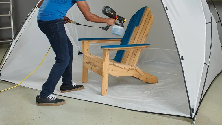 A person using a spray gun on a wooden chair inside a Wagner Spraytech spray shelter with a clear front panel and integrated floor.