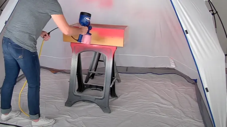 A person using a HomeRight Finish Max paint sprayer on a wooden piece inside a spray shelter, with overspray protection on the floor.