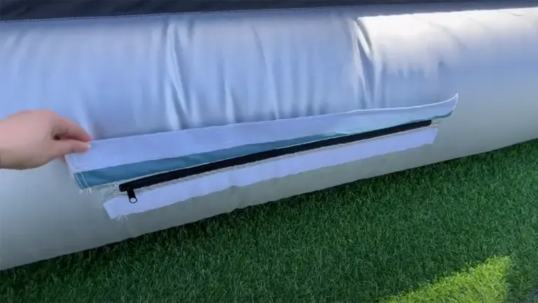 A hand unzipping a section of the white inflatable structure, revealing a black mesh layer underneath, against a backdrop of green artificial turf.