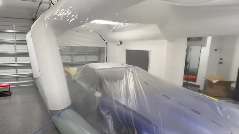 
Inside a white inflatable paint booth with a car covered in protective plastic, featuring the booth's curved structure and a glimpse of a garage setting in the background.