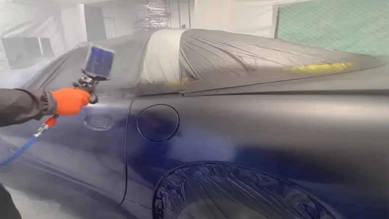 A person’s hand wearing an orange glove is using a paint spray gun on a car inside a white inflatable paint booth, with the car's surface partially covered for protection.