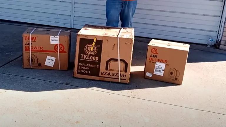Three cardboard boxes on the ground, labeled 'TKLOOP INFLATABLE SPRAY BOOTH' and 'AIR BLOWER', with partial view of a person standing behind them.