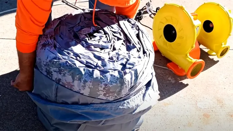 A person in an orange shirt is folding a deflated inflatable structure, with a camouflage pattern, next to two yellow air blowers on the ground.