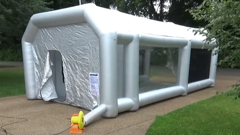 An inflatable paint booth set up outdoors with a reflective silver exterior, clear side windows for visibility, and a large entrance flap, all framed by sturdy inflatable supports. An air filtration fan is visible in the foreground.