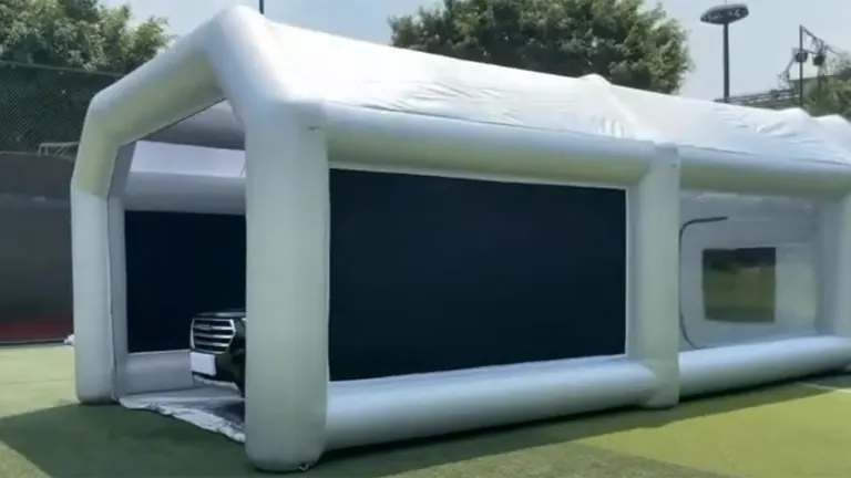 A large inflatable paint booth installed on an outdoor surface, featuring wide, clear PVC windows and an open entrance revealing a car inside, all supported by robust inflatable beams.