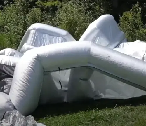 An inflatable paint booth collapsed on the grass, its white beams twisted and the clear plastic walls sagging, in the process of being set up or taken down, amidst a natural green setting.