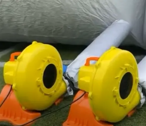Two bright yellow air blowers with orange bases, designed for inflating structures, positioned on the ground next to a partially inflated silver object, possibly a paint booth.