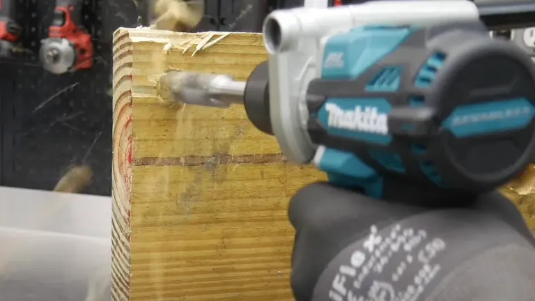 Makita cordless hammer drill in action, drilling into a wooden plank with visible sawdust, in a workshop setting.