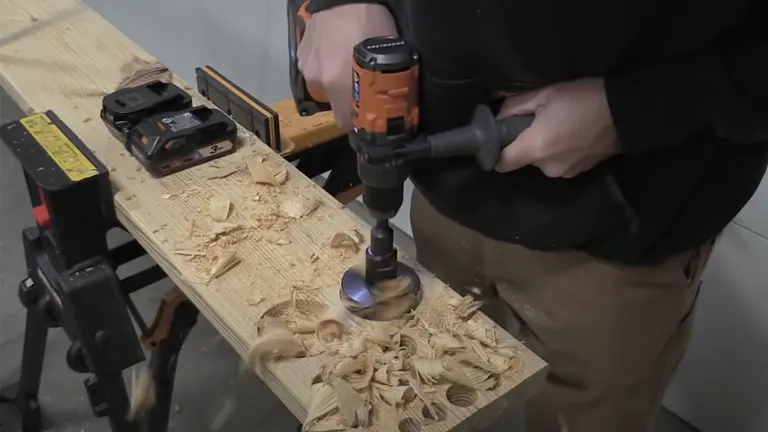 A person operates an orange RIDGID hammer drill, boring into wood with shavings scattered on the workbench.