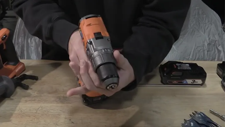 Close-up of hands adjusting the chuck of an orange RIDGID cordless hammer drill, with drill bits and a battery pack nearby.