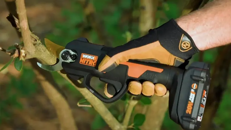 A gloved hand operates a Worx NITRO electric pruning shear, cutting through a thick branch.