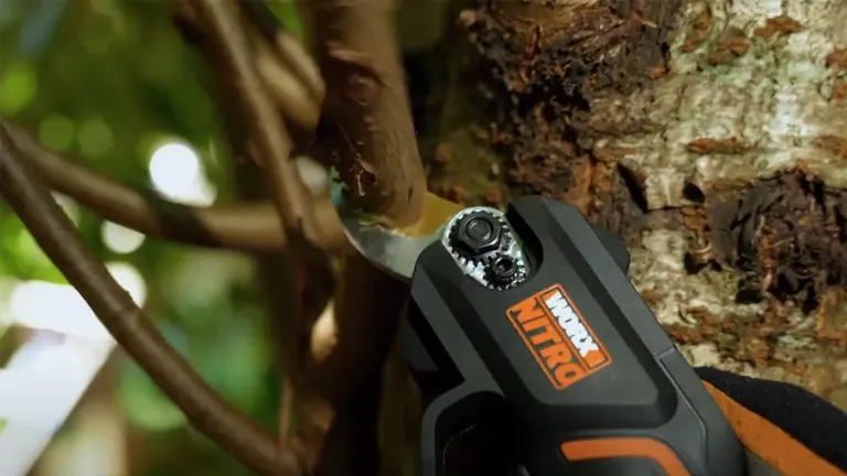 A close-up action shot of a Worx NITRO pruning shear cutting into a brown tree branch, with the focus on the blade and the gear mechanism.