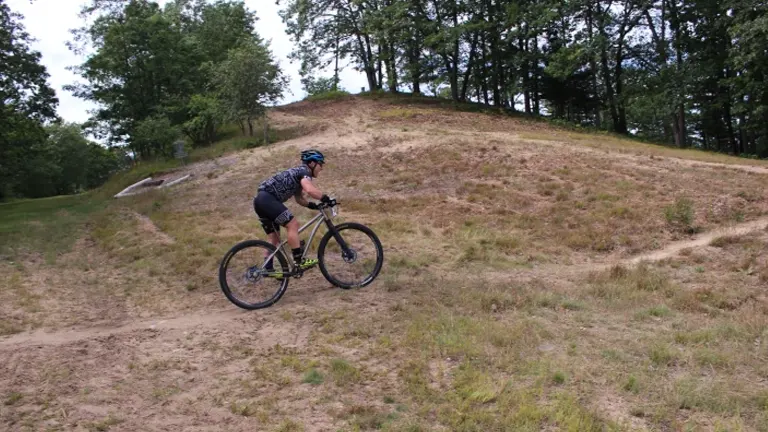 A cyclist in black attire is riding a mountain bike on a sandy trail with a hill and trees in the background.

