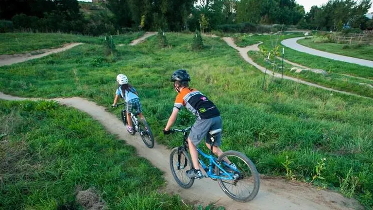 Two cyclists, one in a white helmet and the other in an orange and black outfit, ride on a curving dirt bike path in a grassy area, with a paved path nearby.

