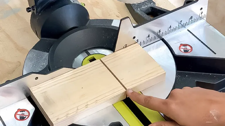 Finger pointing at a measurement mark on a wooden board at a miter saw station, preparing for a cut