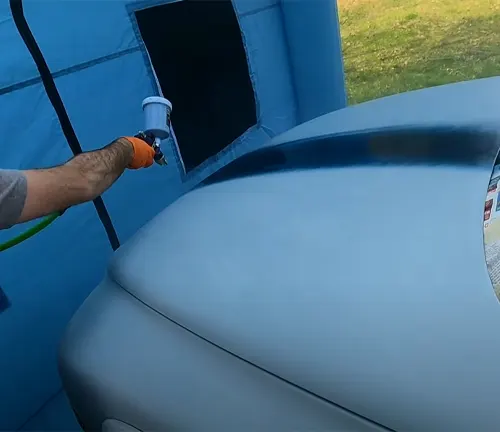 
A close-up of a car being spray painted with a focus on the spray gun and the vehicle's curved hood, inside a blue booth.