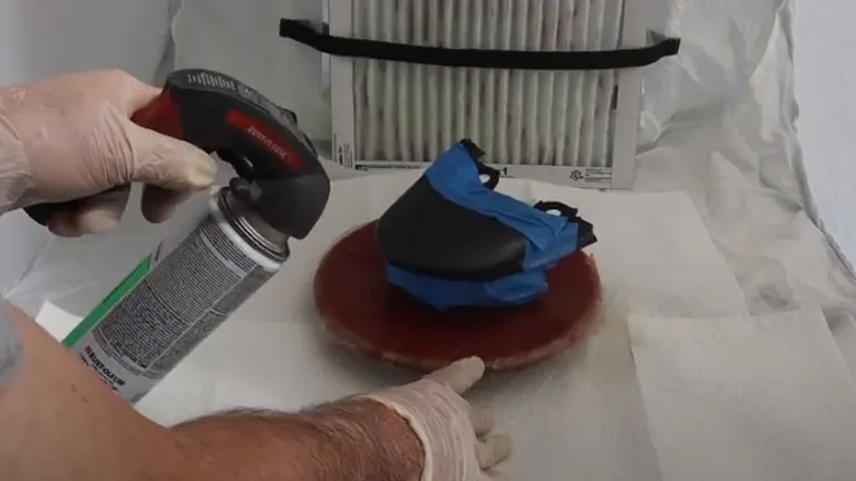 A person in gloves spray painting a masked object on a turntable inside a spray shelter, with a focus on precision and protection.