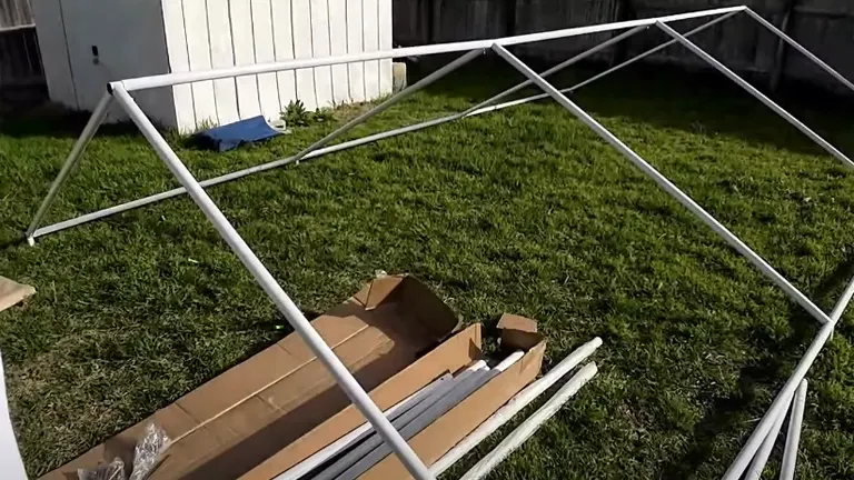 Framework of a portable paint booth under construction, with metal poles laid out on a grassy yard next to an open cardboard box with parts.