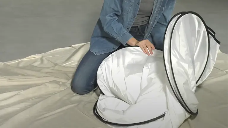 A person in a denim jacket and jeans is in the process of folding a Wagner Spraytech paint spray booth, which is halfway collapsed and resting on a drop cloth.