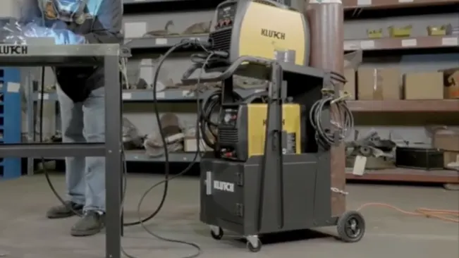 A Klutch 2-Tier Welding Cart equipped with a welder and hoses, set in an industrial workshop.
