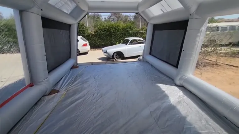 Inside view of an inflatable paint booth with a gray floor, showing the open exit overlooking a parked car and a fenced outdoor area.