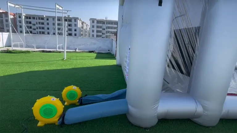Two yellow air blowers connected to a white inflatable paint booth on a grassy field with buildings in the background.