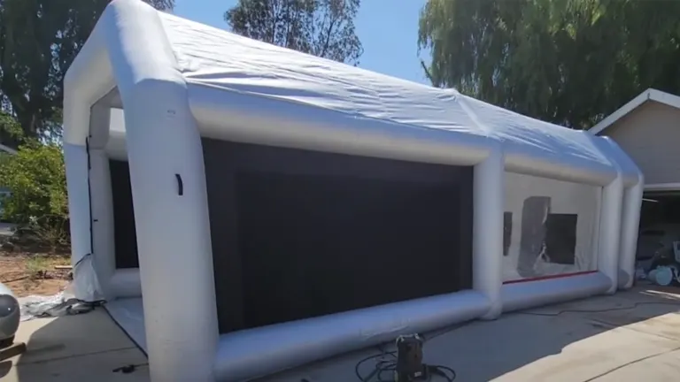 A white inflatable paint booth with a sloped roof and large black mesh windows, situated outdoors next to a residential driveway and garage, with equipment visible on the ground.