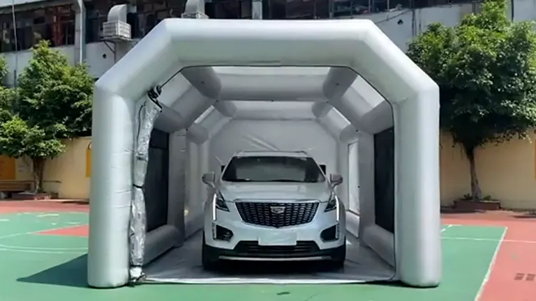 A white inflatable paint booth with a car positioned inside it, set up on a paved area with buildings in the background. The booth has a sturdy structure with a covered top and open ends, allowing easy access for vehicles.