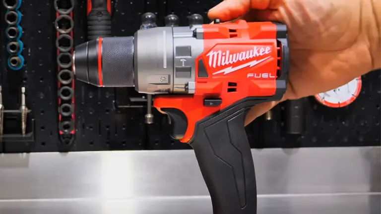 A hand holding a red and black Milwaukee M18 Fuel Hammer Drill/Driver against a backdrop of a well-organized tool wall with various tools hanging neatly.