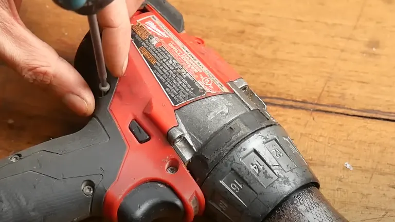 A hand using a screwdriver to service a well-used red and black hammer drill, with a focus on the screws near the drill's label on a wooden surface.